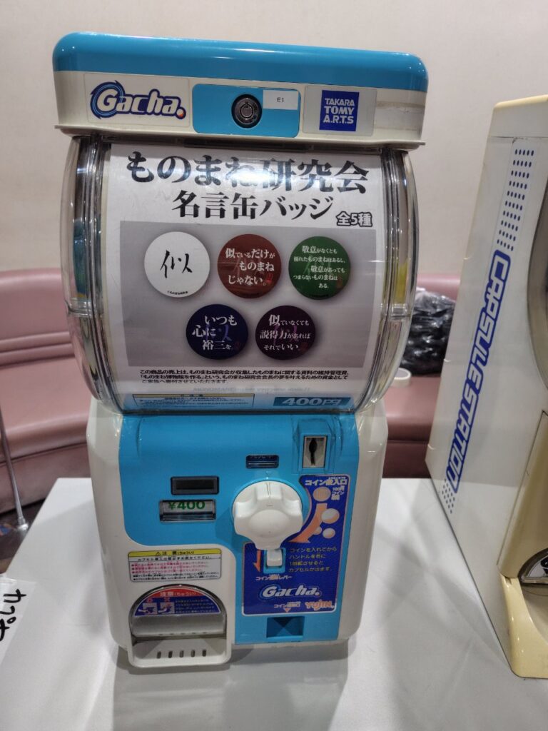 ロビーに設置された「ものまね研究会」オリジナル缶バッジ・ガチャガチャ (レッツゴーよしまさ制作)